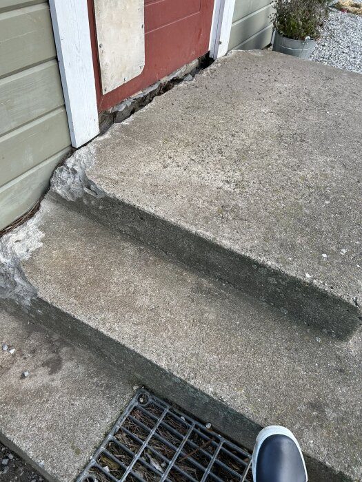 Betongtrappa vid dörr med skador vid mötet mellan fasaden och trappan, synlig röta och reparationsarbete i trä bakom trappan.