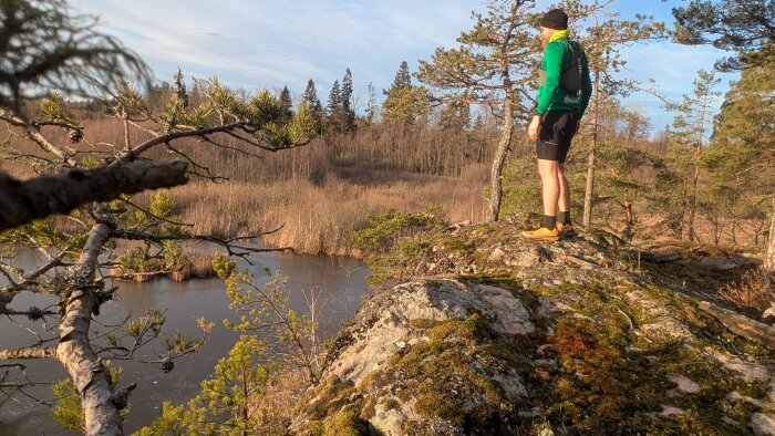 Man står på en klippa med utsikt över en frusen sjö och skog, klädd i grönt och svart med en ryggsäck. Solbelyst scen med barrträd runtom.