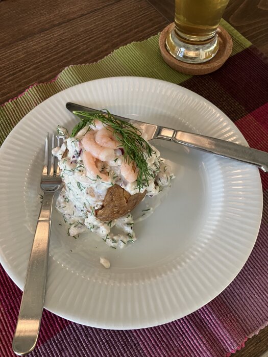 Bakpotatis toppad med skagenröra och räkor, serverad med bestick på vit tallrik. Ett glas dryck i bakgrunden.