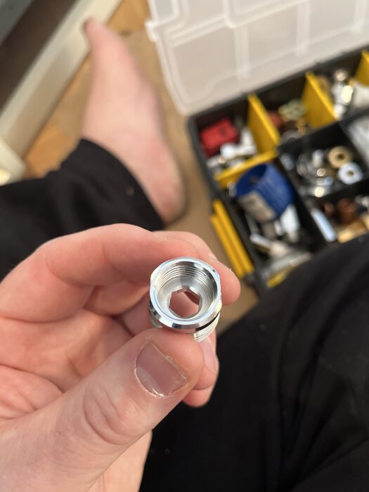 Närbild på en hand som håller en metallanslutning, med en verktygslåda fylld med olika kopplingar synlig i bakgrunden.