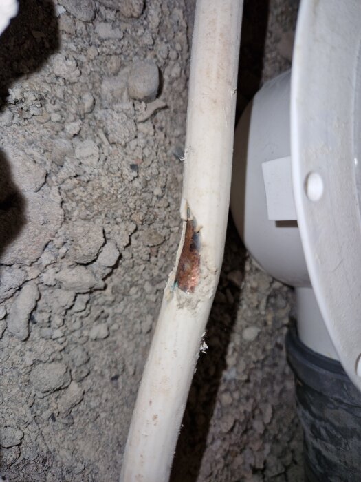 Skadat vitt plaströr med intern kabel synlig och omgivande jord eller murbruk.