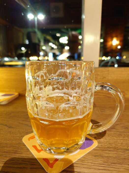 Ett halvtomt ölglas står på ett bord på en ölunderlägg med ljus i bakgrunden.