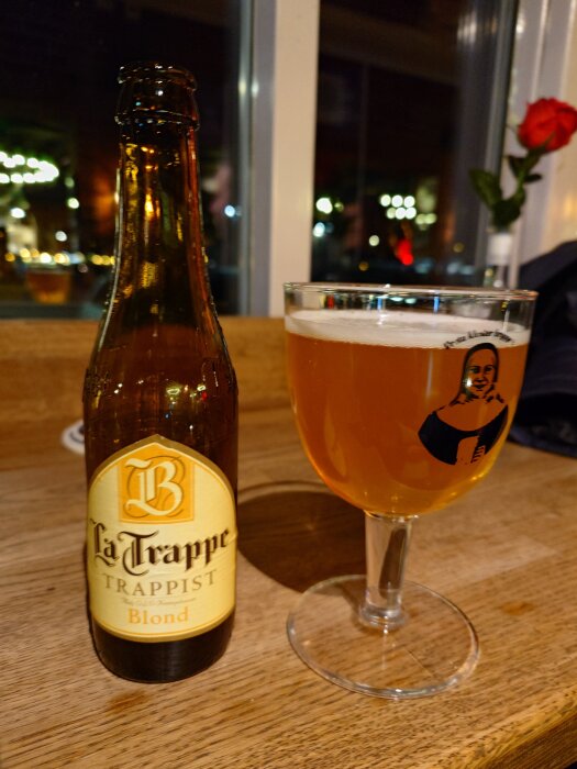 Flaska och glas med La Trappe Trappist Blond öl på ett träbord, ljus belysning och en röd ros i bakgrunden.