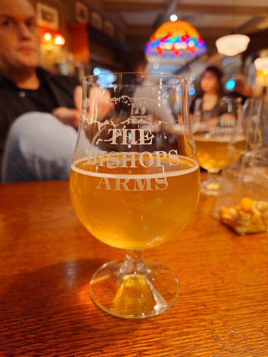 Ett glas öl på ett träbord med texten "The Bishops Arms". Flera suddiga personer syns i bakgrunden på en pub med färgglada lampor.
