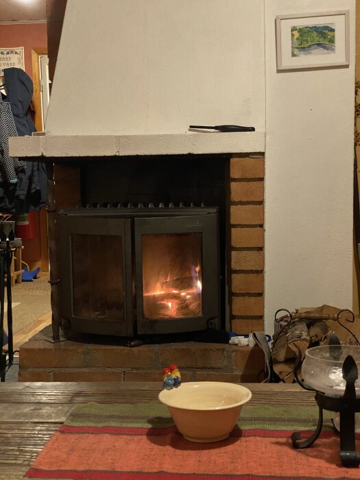 Braskamin med eld, vit puts och tegel. Framför en dukad bordsyta med skål och dekoration. Ved förvarad vid sidan om kaminen.