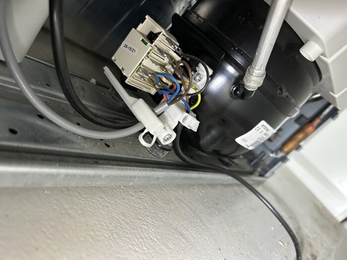 Närbild av kylskåpskompressor med kablage och anslutningar, fokus på en plastkontakt med flera färgade ledningar.