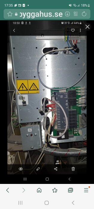 Bild av övertäckt elektronikpanel med kablar och en röd knapp märkt T1 vid en enhet. Varningstekniker och komponentnummer synliga.