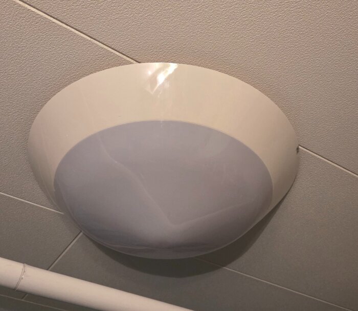 Rund taklampa monterad i ett vitt innertak bredvid ett vitt rör.