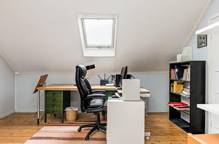 Ett kontor på tredje våningen med datorskrivbord, svart kontorsstol, bokhylla fylld med böcker och ett takfönster som släpper in ljus.