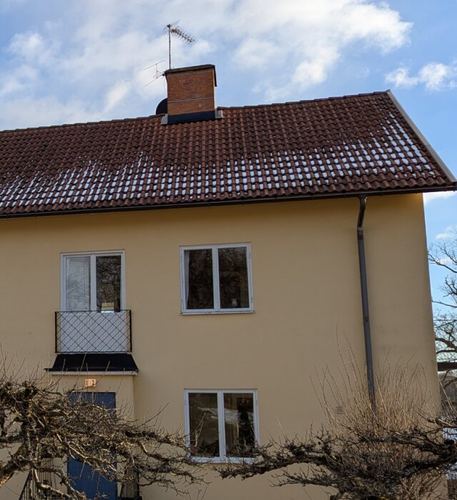 Fasad med gula väggar och brunt tegeltak. Två nya fönster syns på övre våningen, snö på taket. En balkong och dörr på nedre våningen.