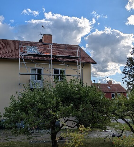 Byggställning framför ett tvåvåningshus under fönsterbyte på tredje våningen. Takfönstret är ersatt och vädret är klart.
