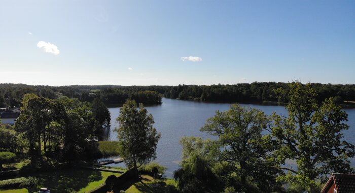 Utsikt över en sjö med omgivande träd och skog under en klarblå himmel, fotad från ett kontorsfönster på tredje våningen.