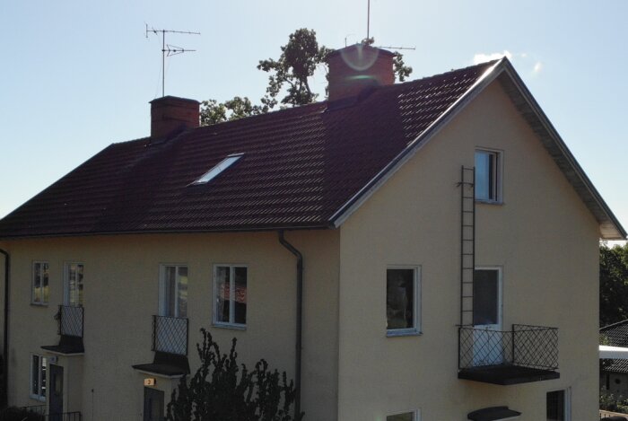 Gult hus med brunt tegeltak, två balkonger, och ett takfönster syns tillsammans med en stege på sidan.