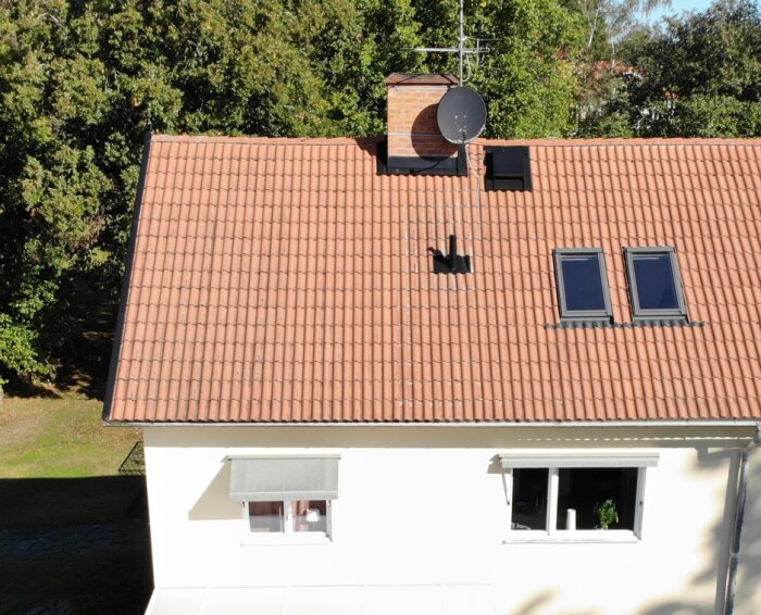 Tak med två nya fönster installerade bredvid en skorsten och parabolantenn, omgivna av gröna träd och blå himmel.