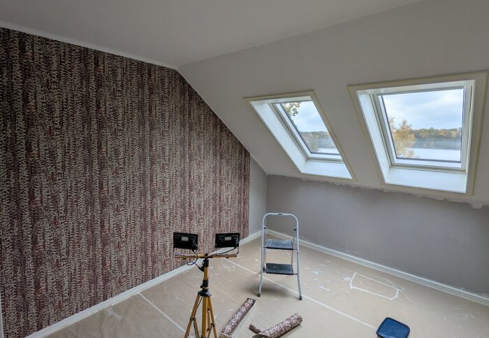 Målat rum med mönstrad tapet, Velux-fönster, hopfällbar stege, arbetslampor och tapetverktyg på golvet under renovering.
