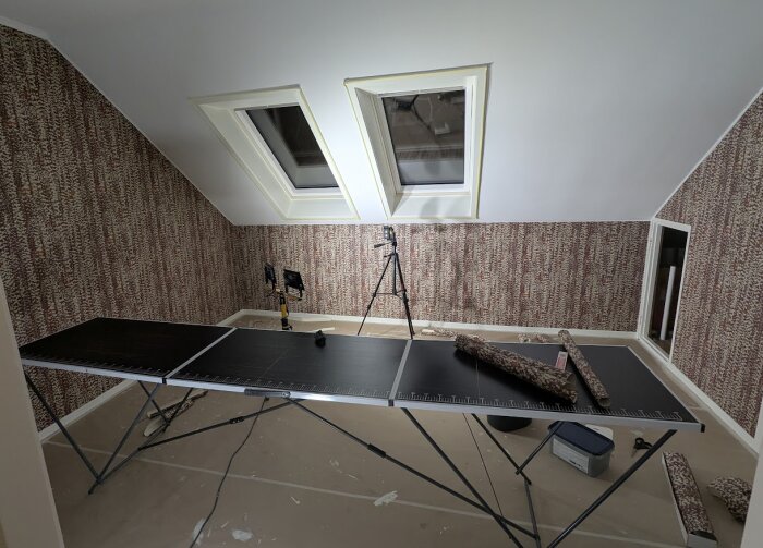 Renoverat kontor med snedtak, Velux-fönster och tapetserade väggar i brunmönstrad tapet. Tapetbord, ljus och utrustning syns också.