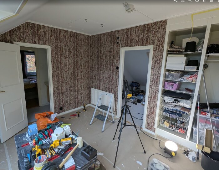 Renovering av kontor med verktyg på bord, ny tapet på väggar och öppna garderober fyllda med föremål. Stege och bygglampor syns i rummet.