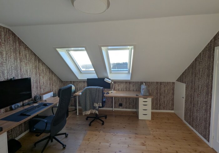 Kontor på tredje våningen efter renovering med träbord, stolar och Velux-takfönster. Träpanelgolv och mönstrade väggtapeter syns tydligt.