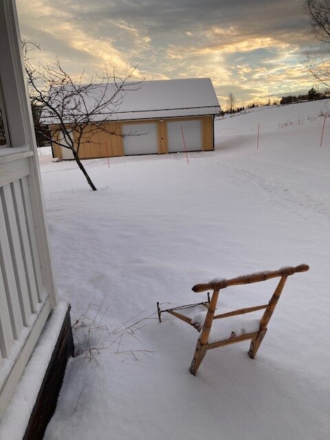 Snölandskap i Brunflo med ett ensamt träd och en snötäckt stol vid ett hus. Solnedgång i bakgrunden.