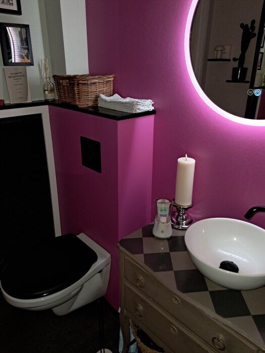 Badrum med rosa vägg, svartvit toalett, vit handfat på gammal byrå, runda spegel med belysning, korg, stearinljus, tandkräm i förpackning.