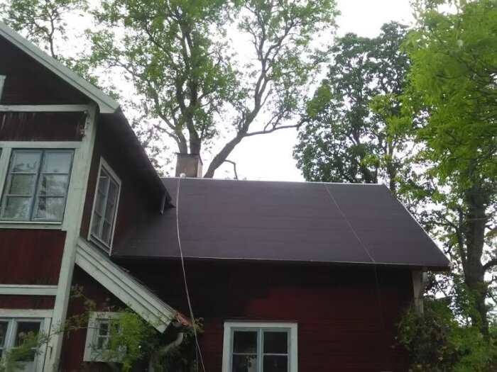 Rött trähus med brant tak och vitmålade fönster, omgiven av träd.