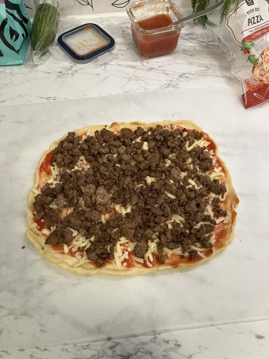 Hemlagad pizza på bakplåtspapper med tomatsås, riven ost och köttfärs. Moulesås i bakgrunden.