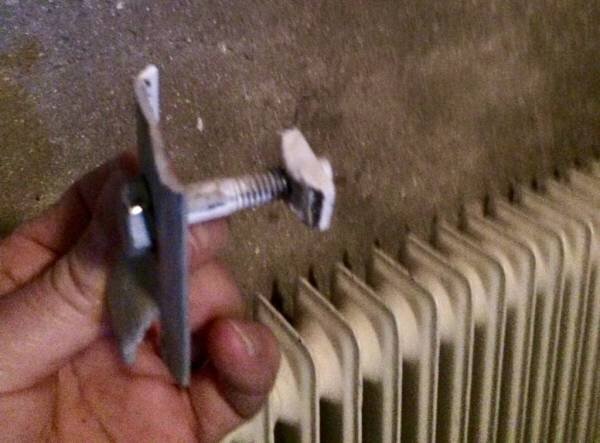 Radiatorfäste med skruv hålls i hand framför en sektionsradiator, redo att tas bort för demontering.