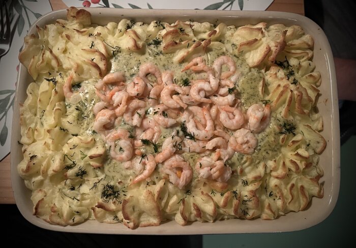 Fiskgratäng med lax, torsk, räkor, hemgjort mos, citron och dillsås i en ugnsform på ett dukat bord.