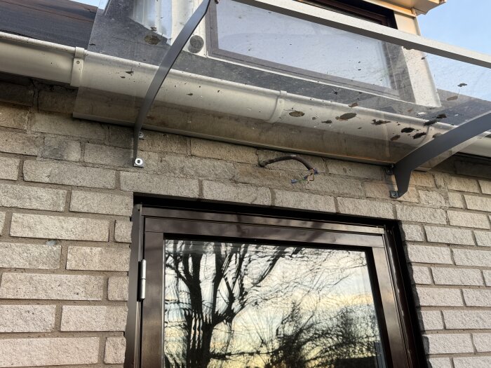Murbruk kring ytterdörr med synlig kabel och skadad murning under balkong, reflektion av träd i glas.