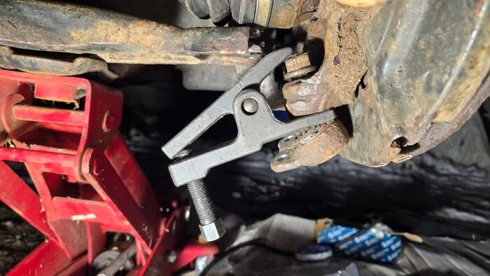 Bilupphängning under reparation med ett avdragarverktyg fäst på rostiga delar vid en V70. En röd domkraft syns till vänster.