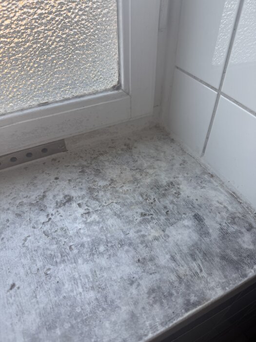 Fönsterbräda med mögel i ett badrum, vit och grå yta med tecken på fukt och tillväxt av mögel nära ett frostat fönster och kakelvägg.