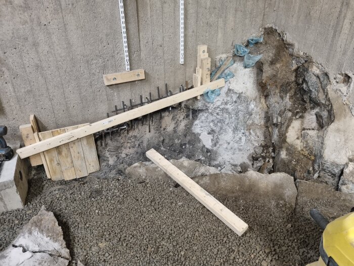 Byggarbetsplats med förstärkning av betongvägg med träbjälkar och armeringsjärn. Grus och berggrund synliga vid foten av väggen.