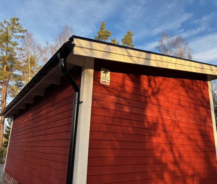 Röd Attefallshusvägg med vita detaljer, svart stuprör och övervakningsskylt under blå himmel.