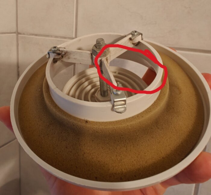 En rund vit föremål med metallskruvar och ventilerande element, handhållen mot kakelvägg.