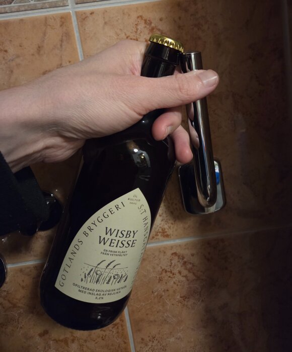 Hand håller i en flaska Wisby Weisse från Gotlands Bryggeri i färd med att öppna den på ett handtag mot en kaklad vägg.