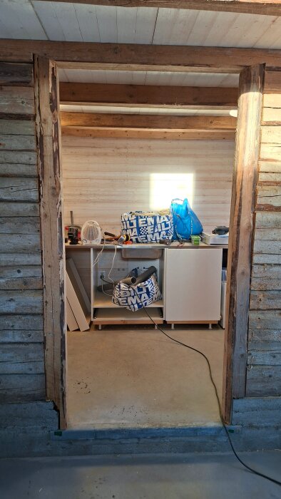 Renoveringsprojekt i träbyggnad, öppning mot rum med verktyg, IKEA-kassar och material på ett skåp, synlig tak- och väggkonstruktion.