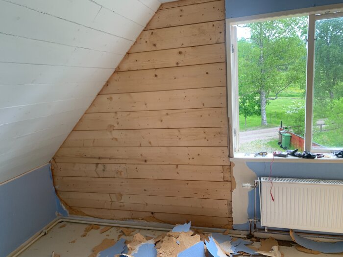 Rum under renovering med träpanel på väggen, pappersrester på golvet och utsikt genom ett öppet fönster mot grönska utomhus.