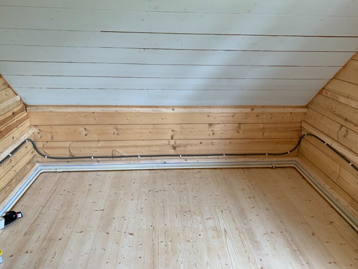 Nymonterat trätak och träpanelväggar i ett renoverat rum med träplankgolv och synliga elkablar längs väggen.