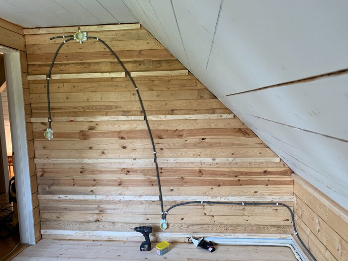 Renoverat rum med träpanel på väggarna och vitmålat snedtak. Elektriska ledningar monterade över träpanel, borrmaskin och verktyg på golvet.