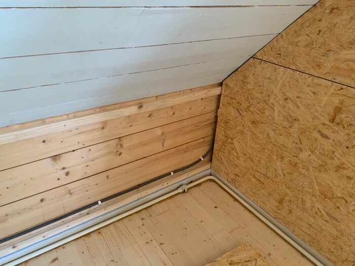 Renoverat rum med träpanel tak och väggar, OSB-skiva på kortvägg, synliga kablar längs golv och vägg, ljust golv.