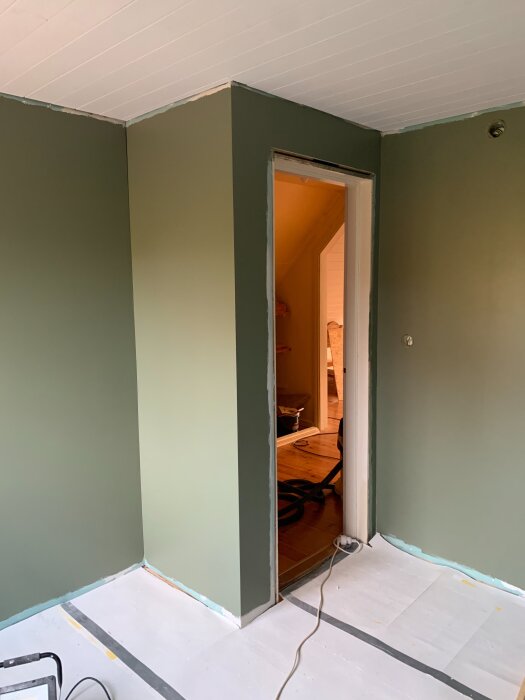 Renoverat rum med gröna väggar, vit takpanel och öppning mot annan del av huset. Golvet är täckt för skydd vid renovering.