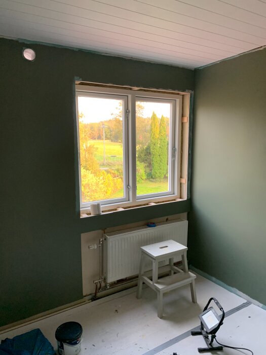 Renoverat rum med gröna väggar, vitmålat tak och nytt fönster. Stegpall, radiator och bygglampa syns intill fönster. Utsikt mot skog.