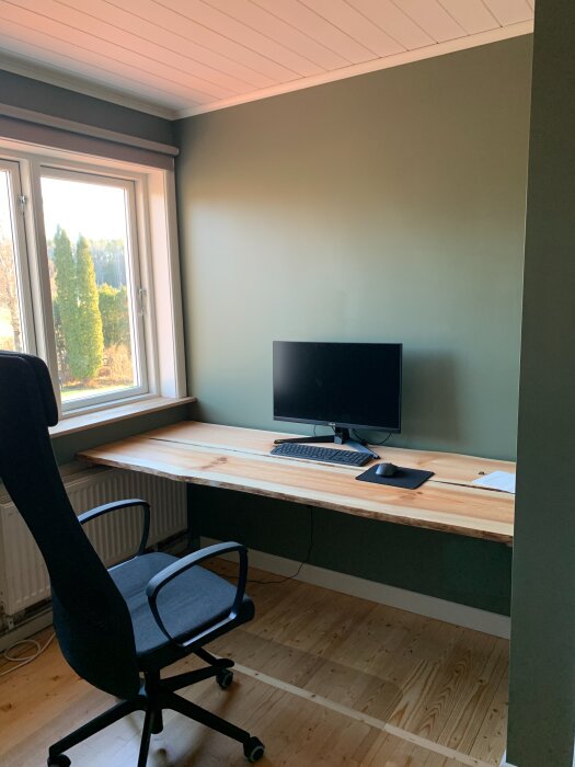 Kontor/gästrum med gröna väggar, nytt skrivbord av furuplankor, datorskärm, tangentbord och stol vid fönster med ljusinsläpp.