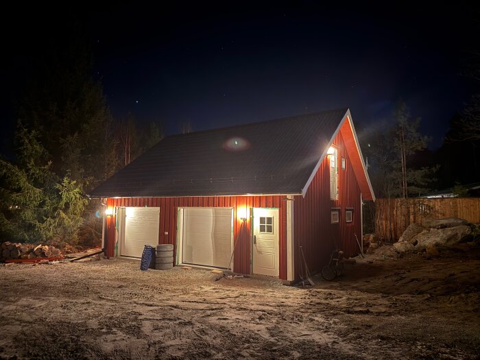 Rött garage med två dörrar och upplyst gårdsplan på kvällen, omgiven av träd och stenar.