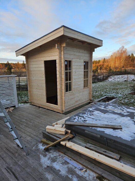 Träbyggnad under konstruktion i vintermiljö med monterade fönster och dörröppning, omgiven av träplankor och snötäckt mark.