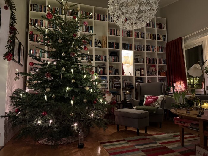 Julgran dekorerad med kulor och ljus i ett vardagsrum med bokhylla i bakgrunden, fåtölj och juldekorationer runtomkring.
