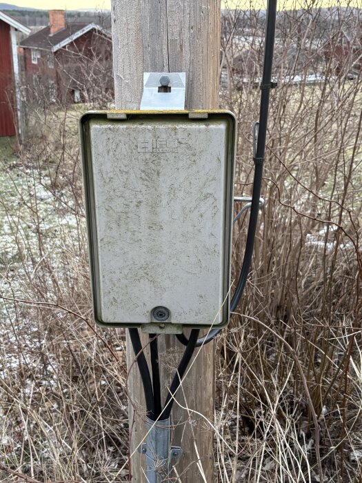 Elektrisk kopplingsbox monterad på trästolpe med kablar. Bakgrund av torra buskar och landskap.