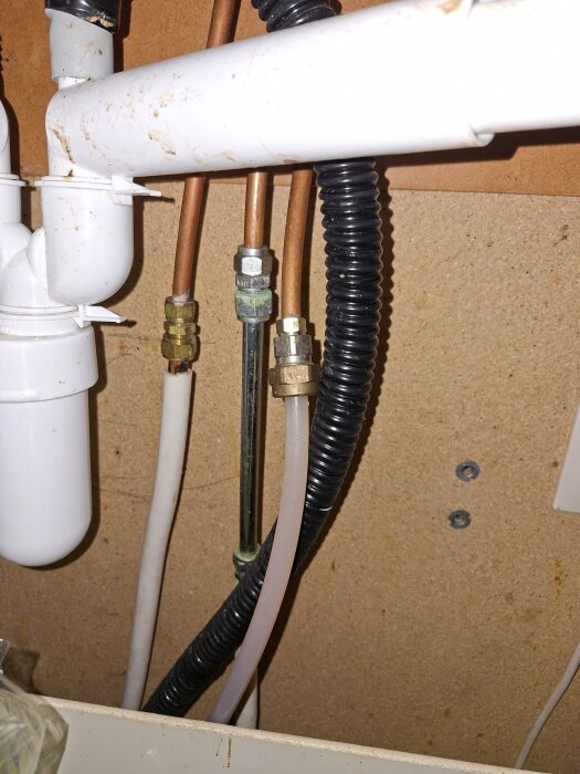 Rörledningar och kopplingar under en köksvask, inklusive plaströr och metallkopplingar, synliga mot en träpanel i bakgrunden.