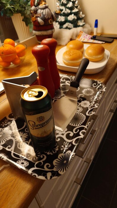 Öppnad burk Staropramen-öl på en bricka med grilltillbehör, bredvid en tomtefigur och en skål med apelsiner på köksbänken.