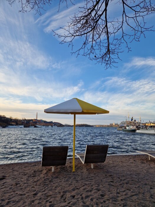Två solstolar under ett gul-vitt parasoll på en sandstrand med utsikt över en sjö och båtar, bar himmel med tunna moln och träd i förgrunden.